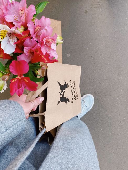 A person holding a floral bouquet featuring pink and white lilies with lush green foliage, wrapped in light brown kraft paper. The bouquet rests on a brown paper shopping bag with black silhouettes of