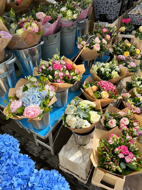 A display of freshly arranged floral bouquets and individual flower bunches at a florist stall, featuring pastel pink roses, white lisianthus, and soft blue hydrangeas wrapped in brown kraft paper and